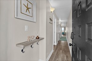 A hallway with a wooden shelf, a starfish decoration, a 'Hello' sign, a vase, and a door with a textured surface. - Beachfront Pet-Friendly 2BR Condo Walkable to Boardwalk (Carolina Beach)
