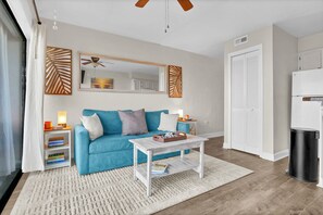 A living room with a blue sofa, a white coffee table, a bookshelf, a ceiling fan, and a white door. - Beachfront Pet-Friendly 2BR Condo Walkable to Boardwalk (Carolina Beach)