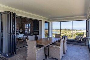 Ein offener Wohnbereich mit Esstisch, Stühlen und einer Couch. Im Hintergrund ist ein Kamin erkennbar, und große Fenster bieten einen Blick auf die Landschaft.