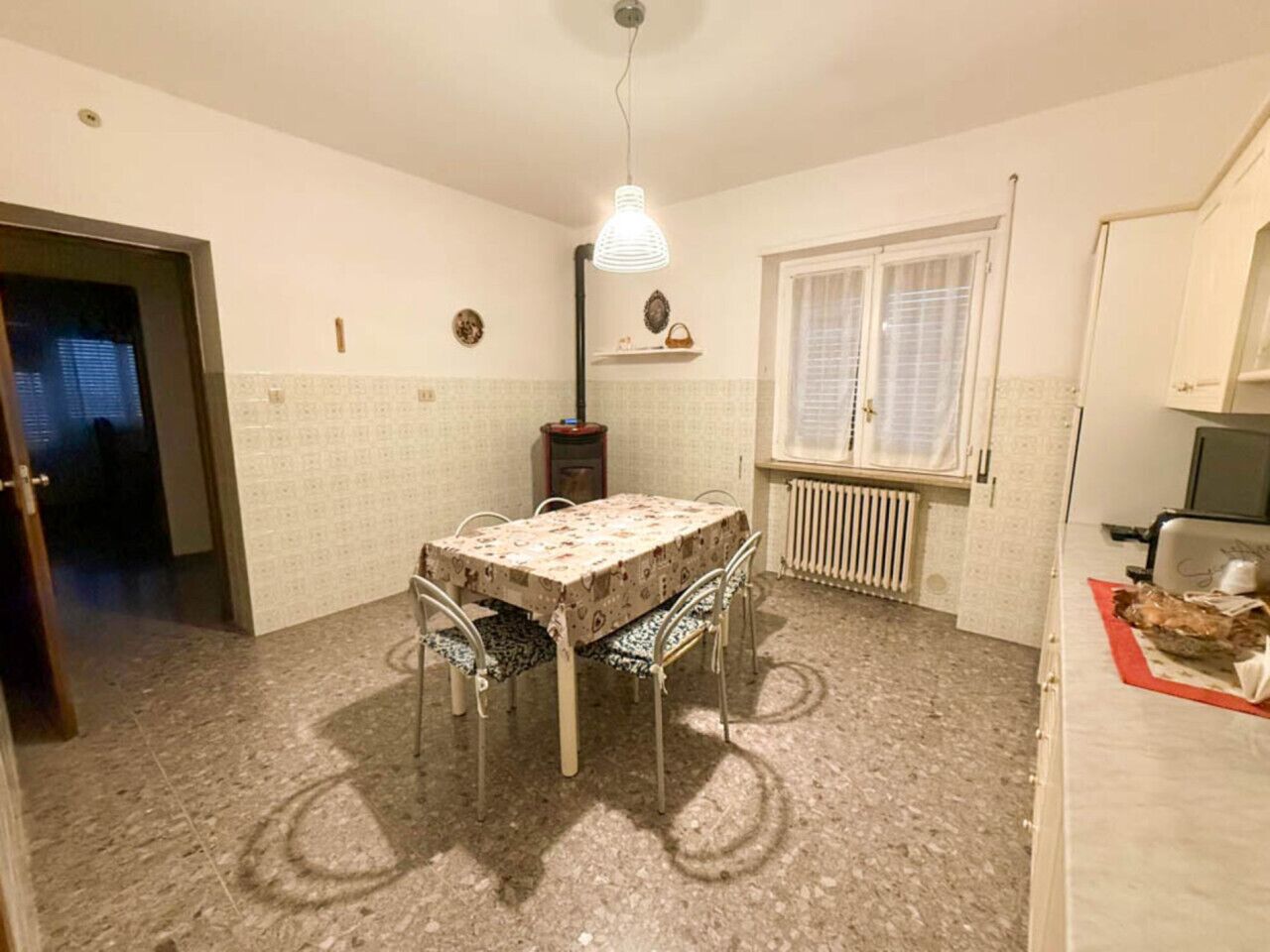 Ein Essbereich mit Tisch und Stühlen, einem Heizkörper, einem Fenster mit Jalousien und einer Tür zu einem anderen Raum.