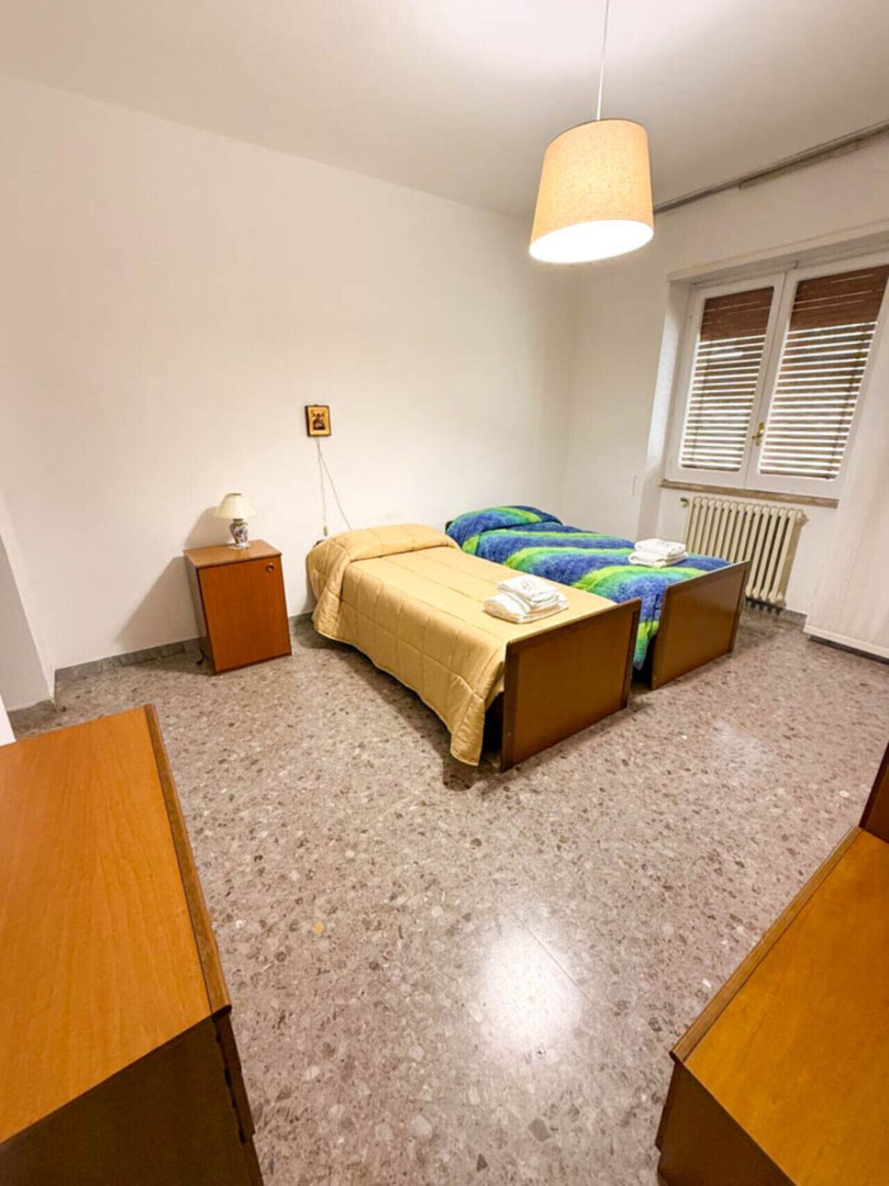 A bedroom with a bed, a bedside table, a lamp, a radiator, and a window with blinds.