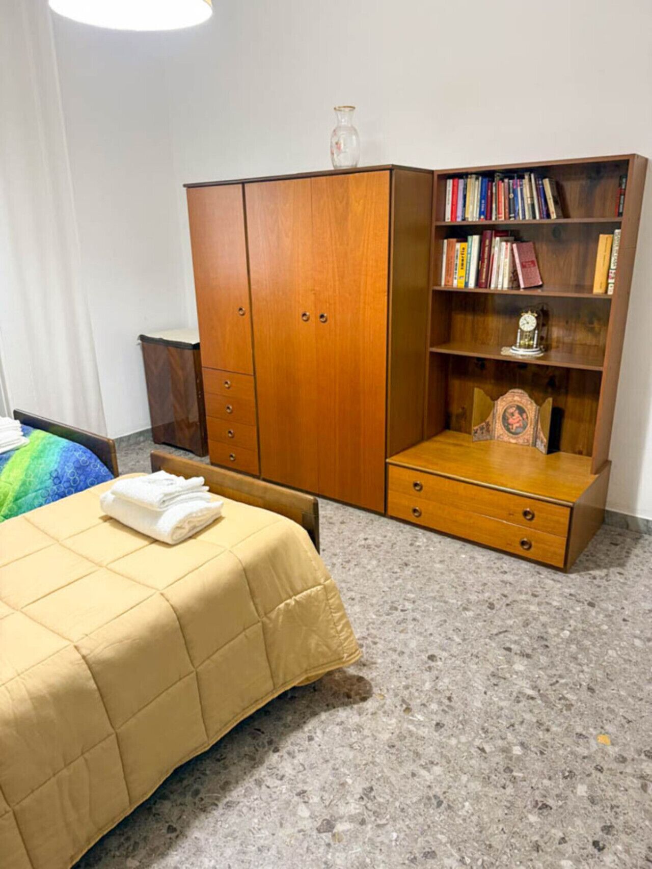 Ein Schlafzimmer mit einem Bett, einem Holzschrank, einem Bücherregal und einem kleinen Tisch mit einer Uhr.
