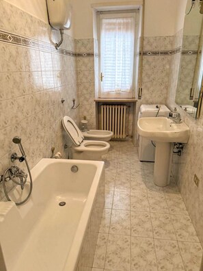 A bathroom with a bathtub, two toilets, a sink, and a window with a curtain. - Casa Vacanze da Francesco (Lettopalena)