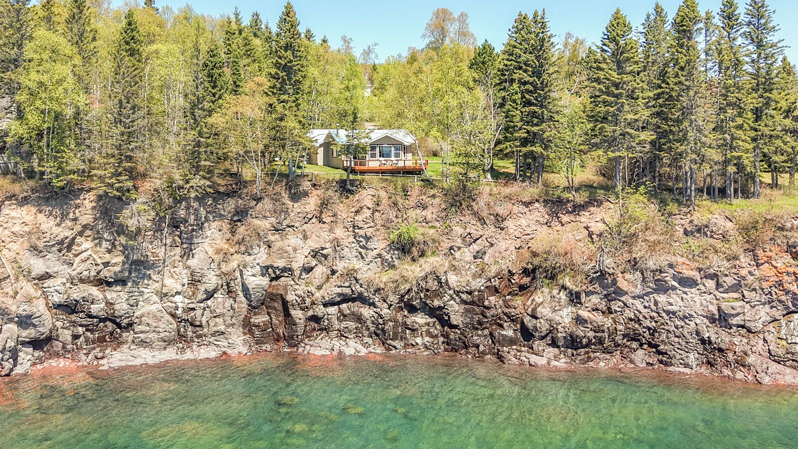 Waterfront property nestled atop rugged cliffs overlooking crystal-clear lake waters, surrounded by towering evergreens and seasonal foliage, offering a secluded and scenic lakeside retreat.