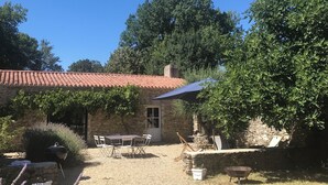 A stone building with a tiled roof, a table with chairs, a grill, and a wheelbarrow.
