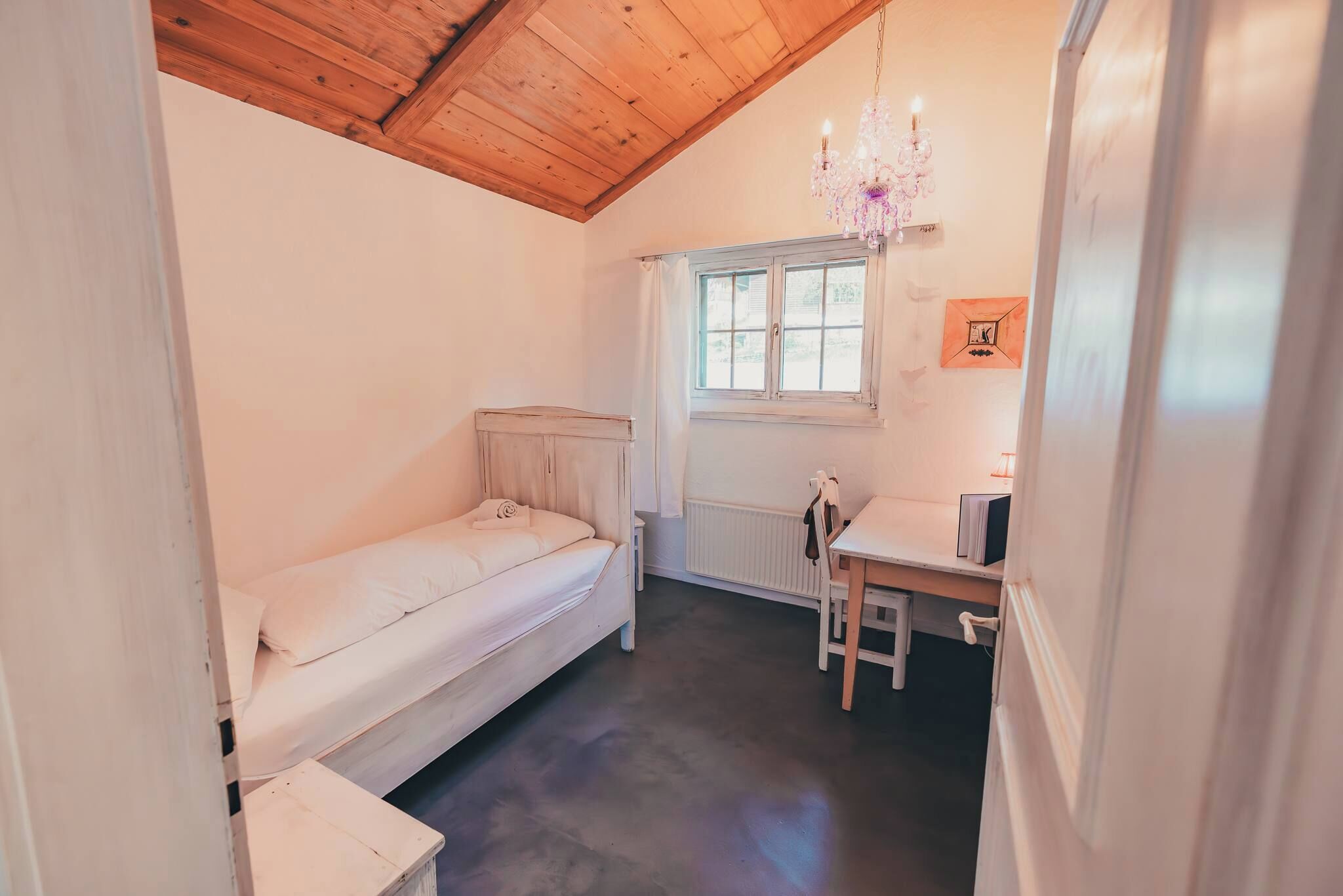 A small, cozy room with a bed, a desk, a chair, a radiator, and a window.