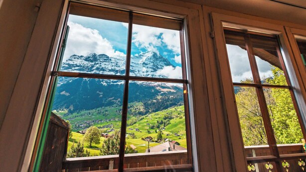 Una ventana con vistas a un paisaje montañoso, que incluye picos nevados y un valle con casas.