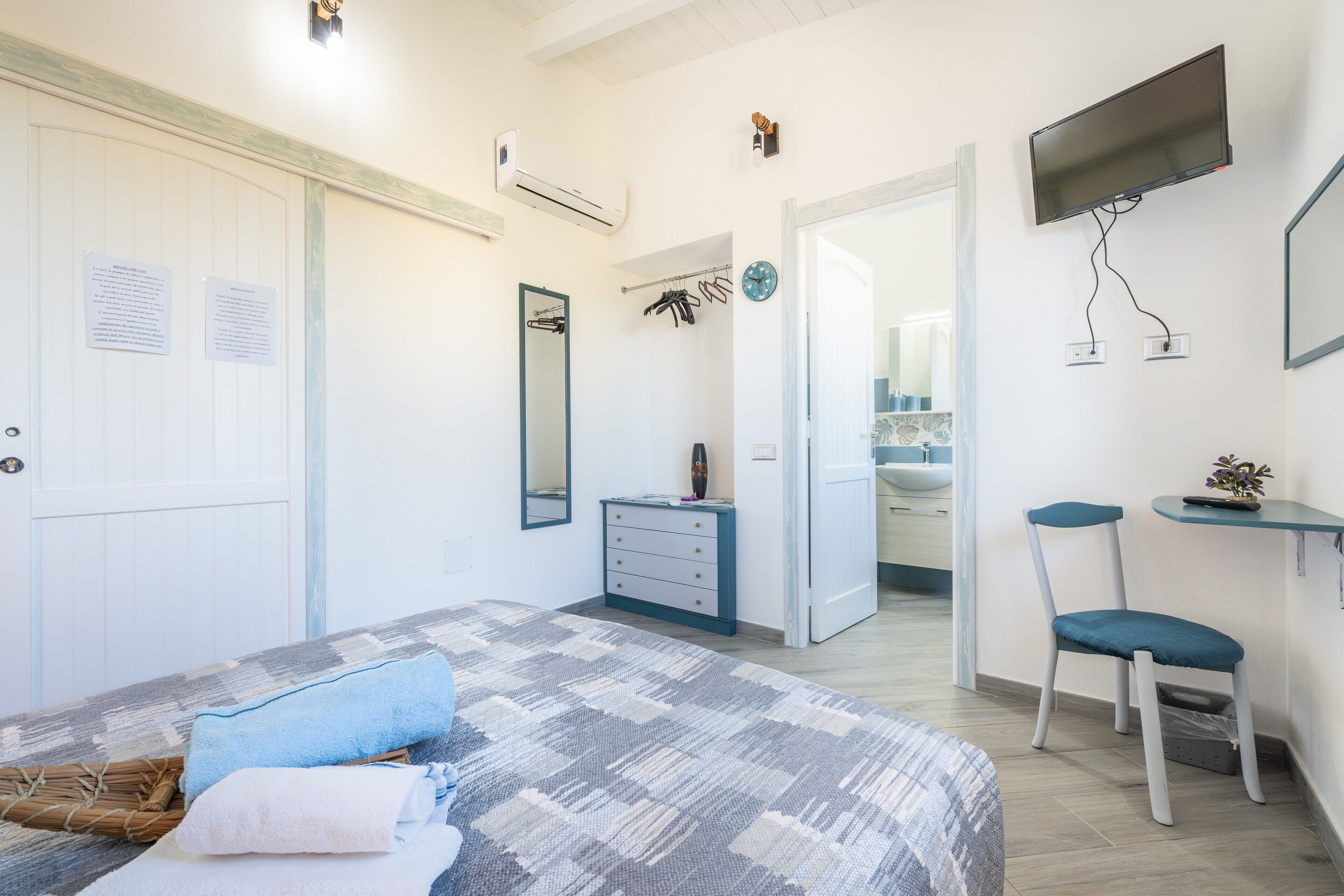 Una camera da letto con un letto, una cassettiera, una sedia, un televisore e un bagno visibile attraverso una porta aperta.