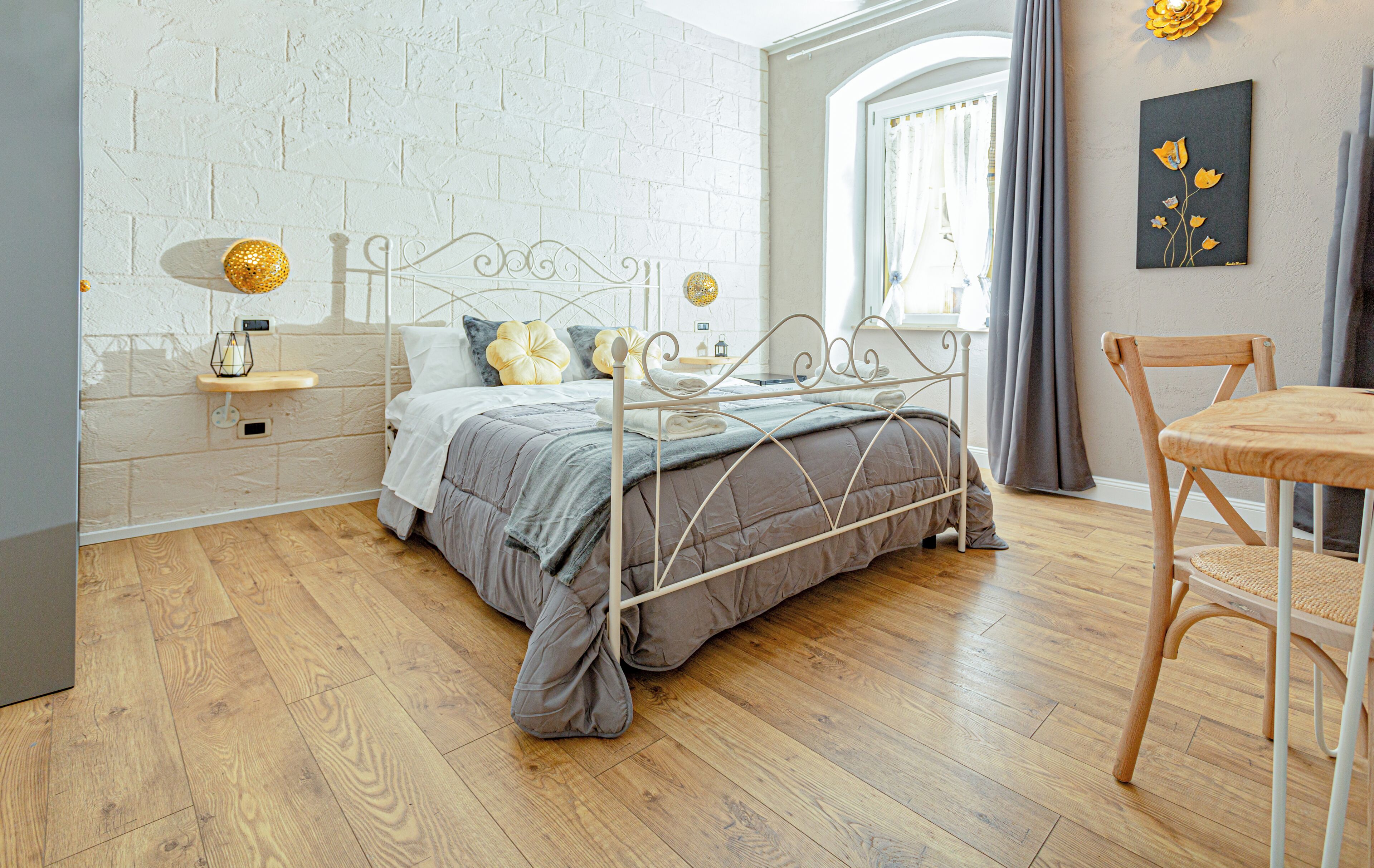 Un dormitorio con estructura de cama metálica, suelo de madera, una mesita de noche con lámpara y una silla.
