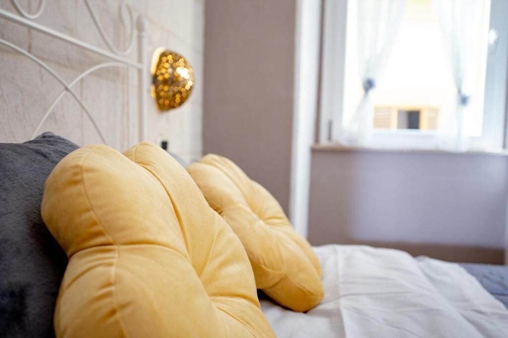 A bed with a white sheet and two pillows, one yellow and one gray, in a room with a white headboard and a window with curtains.