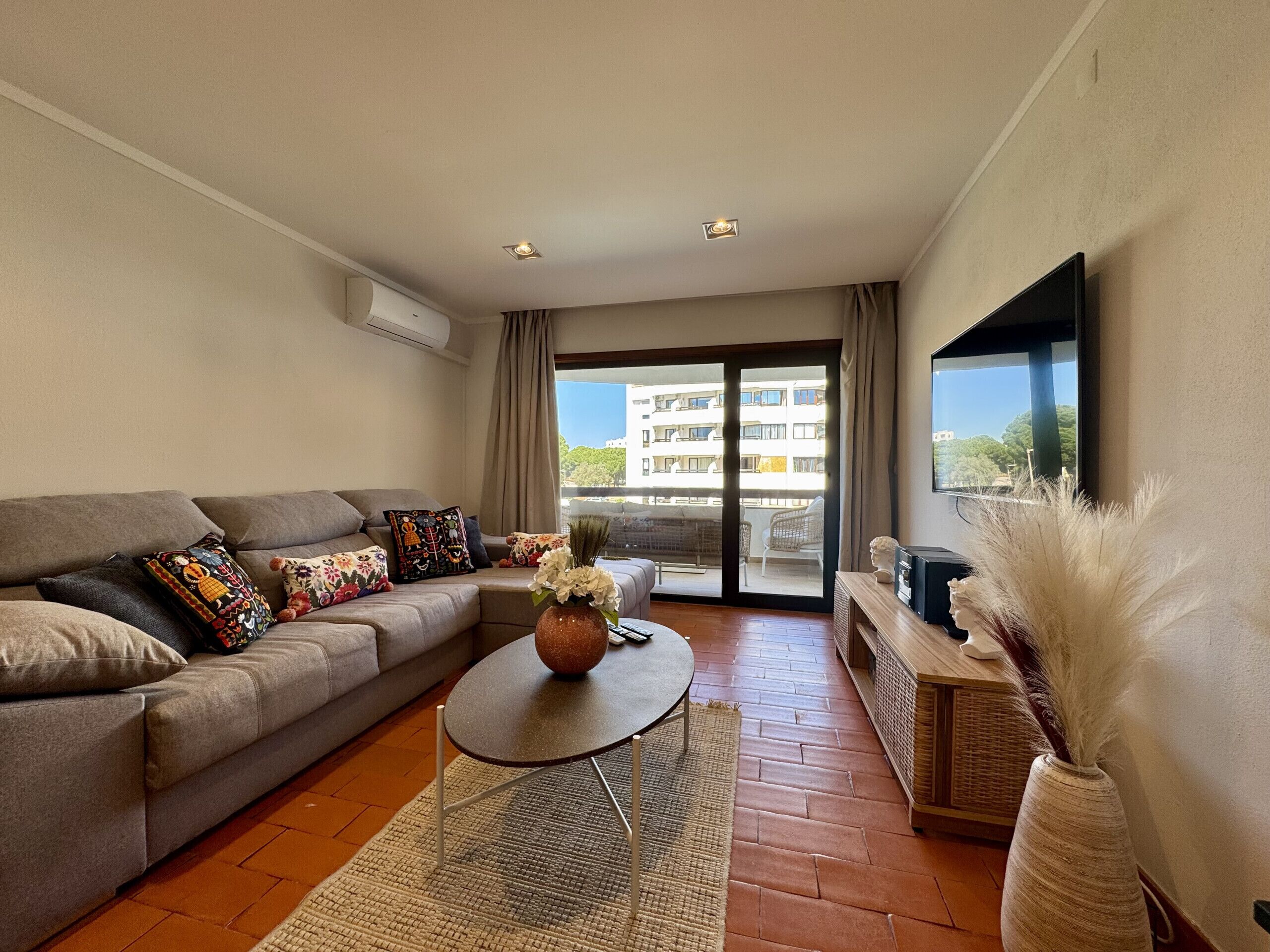 A modern living room with a sofa, a coffee table, a flat-screen TV, and a large window with a view of buildings.