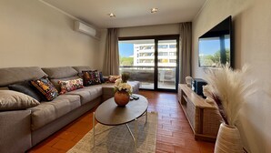 Een moderne woonkamer met een bank, salontafel, flatscreen-tv en een groot raam met uitzicht op gebouwen.