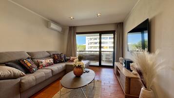A modern living room with a sofa, a coffee table, a flat-screen TV, and a large window with a view of buildings.