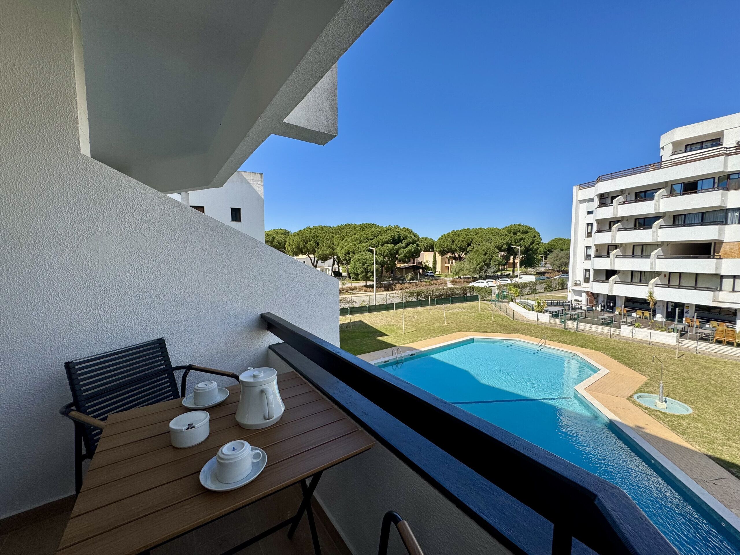 Un balcón con una mesa lista para el té, una piscina y vistas de edificios y zonas verdes circundantes.