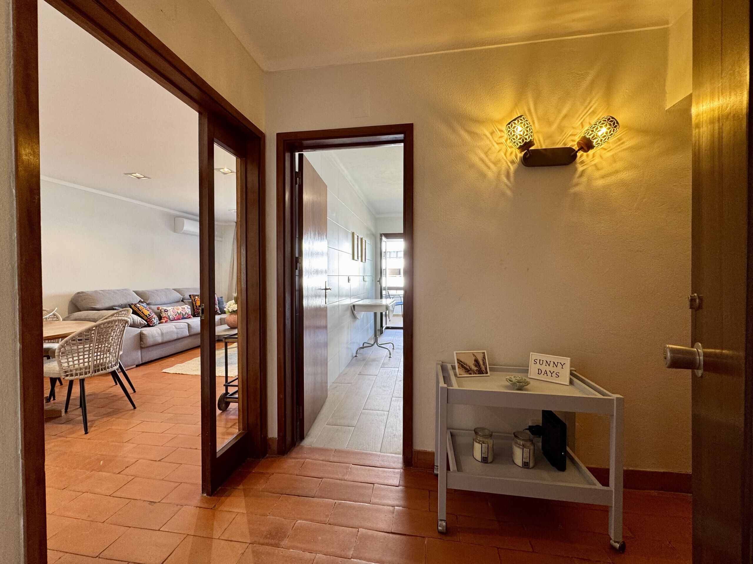 A hallway with a white shelf, a door, and a view into a living room with a sofa and a dining area.