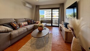 A modern living room with a sofa, a coffee table, a flat-screen TV, and a large window with a view of buildings. - Vilamoura Charming 2 With Pool by Homing (Loulé)