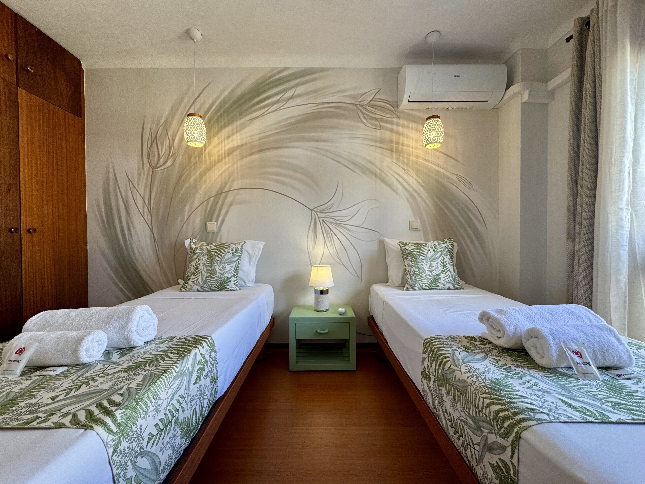 Une chambre d’hôtel avec deux lits, une table de chevet verte, une fresque murale et un appareil de climatisation.