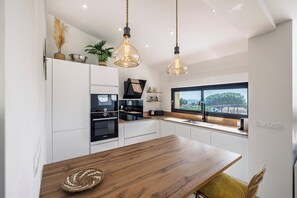 A modern kitchen with white cabinets, a wooden dining table, hanging light fixtures, and a window with a view. - Magnificent villa with sea view, heated swimming pool (optional), pétanque and table tennis. (Roquebrune-sur-Argens)
