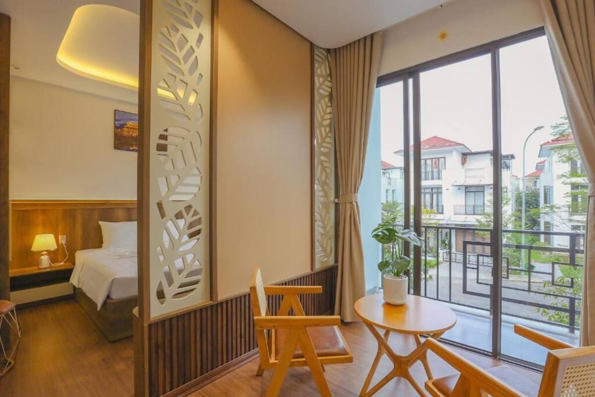 A hotel room with a bed, a desk, a chair, a table, a potted plant, and a view of buildings through large windows.