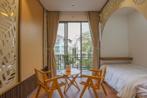 A hotel room with a bed, a round table with chairs, a potted plant, and a view of buildings through a large window. - NGOC THIEN MINH HOMESTAY (Hue)