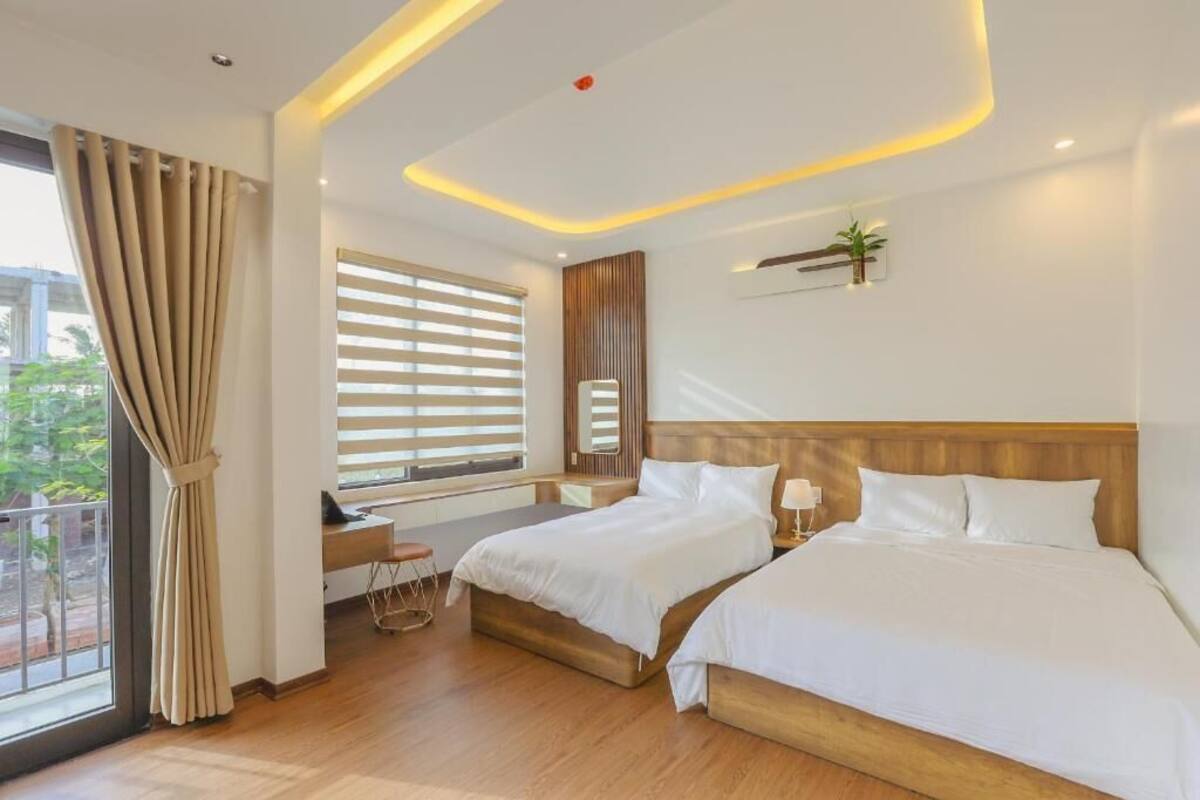A hotel room with two beds, a desk, a window with blinds, and a view of greenery outside.