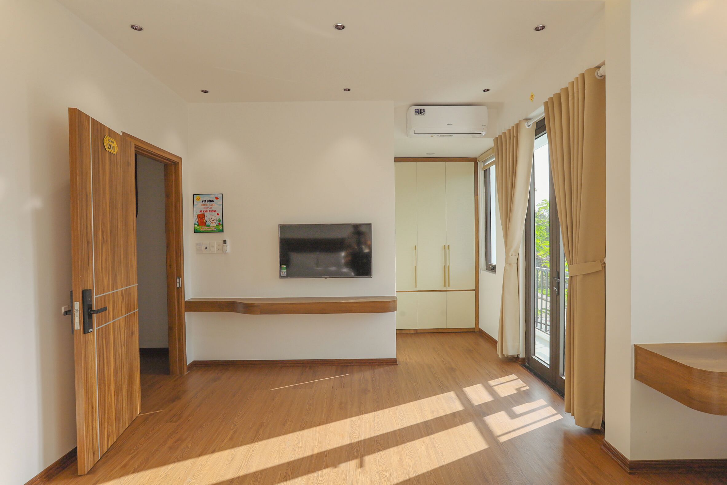 Een hotelkamer met een houten vloer, een flatscreen televisie aan de muur bevestigd, een inbouwbank, een glazen schuifdeur met gordijnen en een airconditioningsysteem.