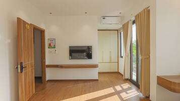 Een hotelkamer met een houten vloer, een flatscreen televisie aan de muur bevestigd, een inbouwbank, een glazen schuifdeur met gordijnen en een airconditioningsysteem.