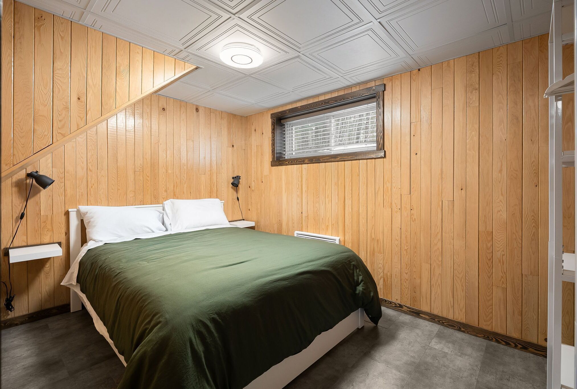 A bedroom with wooden panel walls, a bed with a green bedspread, a bedside table with a lamp, and a window with blinds.