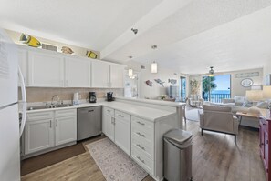 A modern kitchen with white cabinets, a central island, and a view to the living area. - 3BR Gulf View Condo | Steps to Beach + Community Pool | Romar Place 202 (Orange Beach)