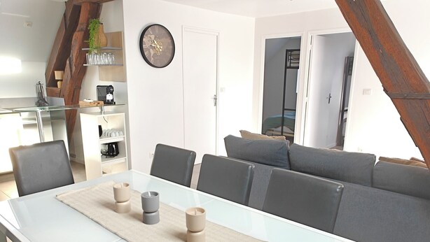 A dining area with a white table and chairs, a clock on the wall, and a lofted space with exposed wooden beams.
