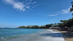 A beach with clear blue water, a sandy shore, and a line of buildings with palm trees. - Spacious 5-bedroom villa/AC at beach resort
15 restaurants/bars. 
All Inclusive! (Cofresi)