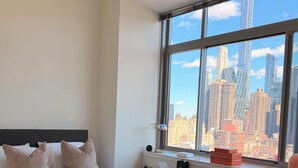 A modern hotel room with a large bed, bedside tables, a radiator, and a window with a city skyline view. - Luxury NYC Penthouse with Amazing Views (New York)