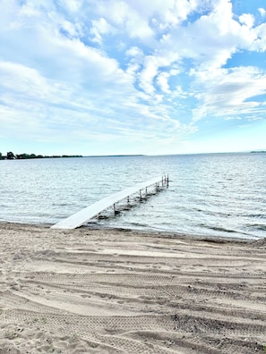 Ein Strand mit einem hölzernen Steg, der sich ins Wasser erstreckt, feiner Sand und ein klarer Himmel mit Wolken.