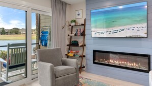 A living room with a fireplace, a large TV, a bookshelf, and a view of a beach through a sliding door. - Coastal Escape with Lake Views & Beach Days Just Minutes Away (Miramar Beach)