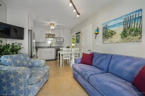 A living room with a blue sofa, a TV mounted on the wall, and a painting of a beach scene. - 2-bedroom apartment in charming Ogunquit steps from the beach with AC and pool (Ogunquit)