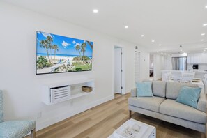 A modern living room with a flat-screen TV, a sofa, a coffee table, and a kitchen area in the background. - Gulf-Front 3BR Heated Pool, Hot Tub, Beach & Balcony (Indian Shores)