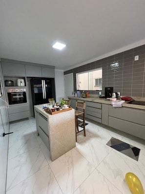 A modern kitchen with a central island, built-in appliances, and a large window. - House in São Pedro da Aldeia - Daily and Seasonal Rentals (São Pedro da Aldeia)