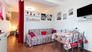 Un espace de vie compact comprenant un canapé, une table à manger et des chaises, un téléviseur à écran plat et des étagères murales ornées d’objets décoratifs.