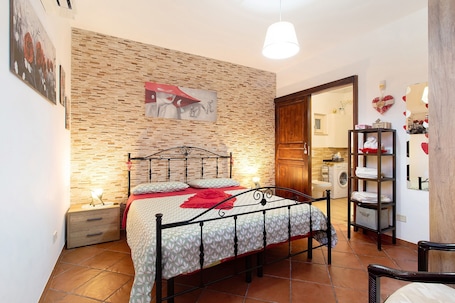 A bedroom with a brick-patterned wall, a bed with a metal frame, a nightstand, a wooden chair, and a shelf with folded clothes.