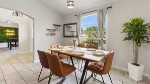 Un comedor con una mesa dispuesta para cuatro, sillas, una lámpara colgante, una ventana con cortinas y una planta en maceta.