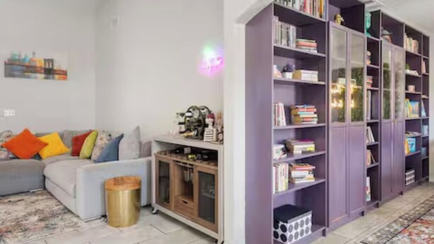 Una sala de estar con un sofá gris, cojines coloridos, una estantería morada y un tapete estampado.