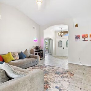 Un salon avec un canapé, une table basse et un tapis. Un couloir mène à une salle à manger agrémentée d’un lustre.