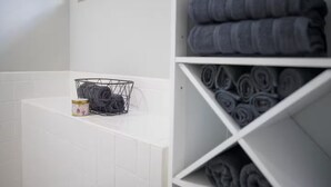 Un baño con paredes de azulejos blancos, una ventana, un perchero con toallas dobladas y un estante con toallas enrolladas.