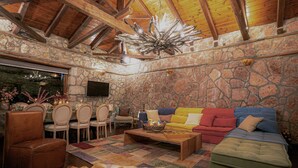 Um quarto com paredes de pedra, teto de madeira, televisão na parede, área de refeições com cadeiras e mesa, e um sofá colorido.