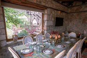 A dining table set with plates, glasses, and candles, surrounded by stone walls and a window overlooking a garden.