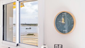 Une pièce avec une fenêtre donnant sur une plage, une horloge fixée au mur et une porte sur laquelle est posé un objet jaune.