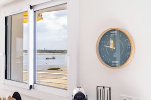 Ein Zimmer mit Fensterblick auf einen Strand, einer Wanduhr und einer Tür mit einem gelben Gegenstand.