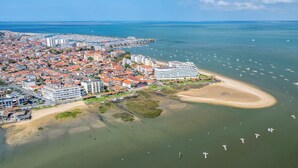 Vue aérienne d’une ville côtière, avec un hôtel imposant, une plage de sable et un port de plaisance rempli de bateaux.