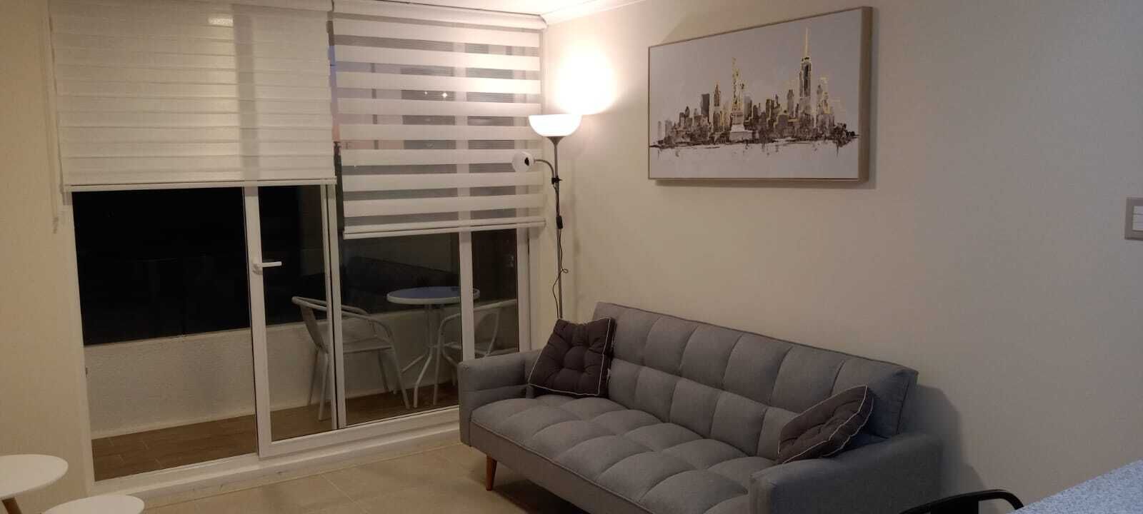 A living room with a grey sofa, a table with chairs, a large window with blinds, and a cityscape painting on the wall.