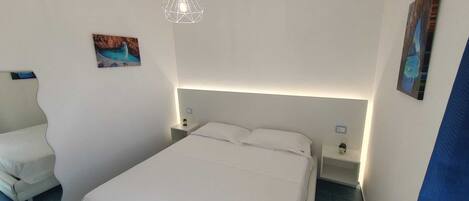 Une chambre d’hôtel comprenant un lit, des tables de chevet, une petite table, une lampe fixée au mur et deux tableaux.
